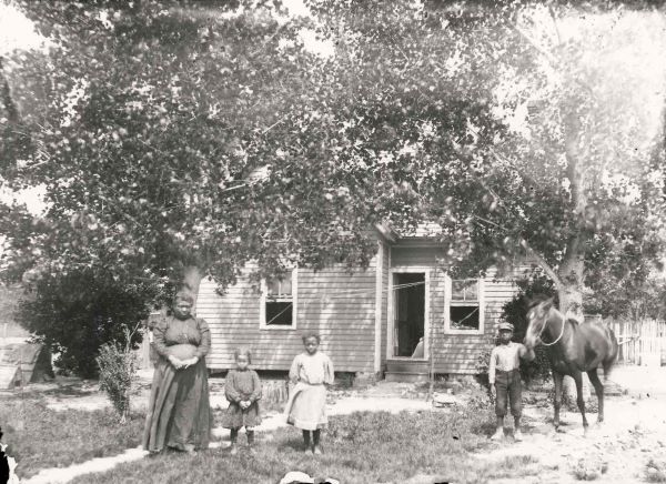 Family standing outside a house. A boy holds the reins of a horse.